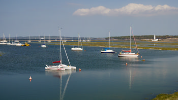 Sailing boats This landscape photograph was taken in the afternoon during late spring and shows several sailing boats and other transport vessels moored on the water at Milford Haven, located in Dorset, United Kingdom. In the distance, Hurst Point Lighthouse is visible on the coastline, providing a recognizable maritime landmark. The calm water reflects the boats, and the shoreline in the background is lined with greenery. The image captures transport on the water, with boats being the central subject against a clear sky and scattered clouds.